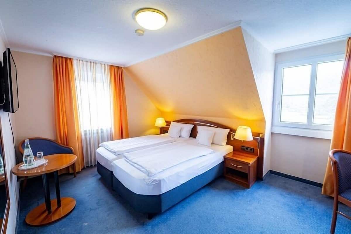 Una habitación de hotel con una cama grande, dos mesitas de noche con lámparas, una mesita pequeña con una botella y vasos, y un televisor en la pared.