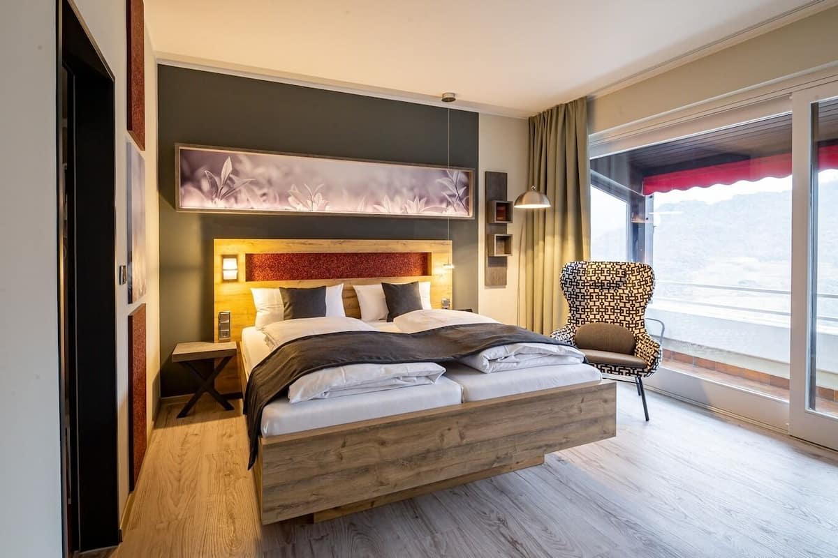 Habitación de hotel moderna con una cama grande, cabecera de madera, una silla y vistas a las montañas a través de la ventana.