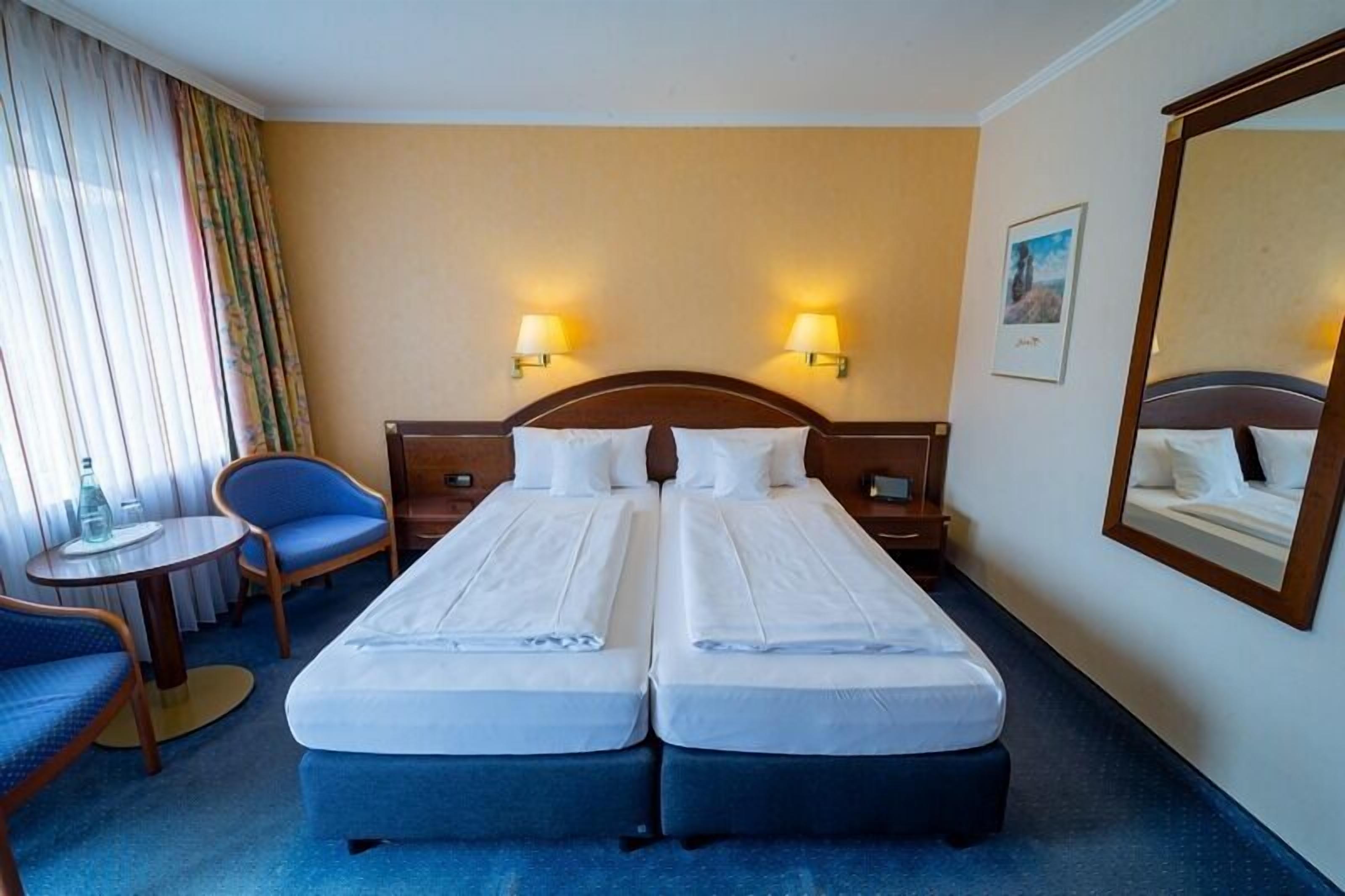 A hotel room with two single beds, a small table with a chair, a large mirror, and two wall-mounted lamps.