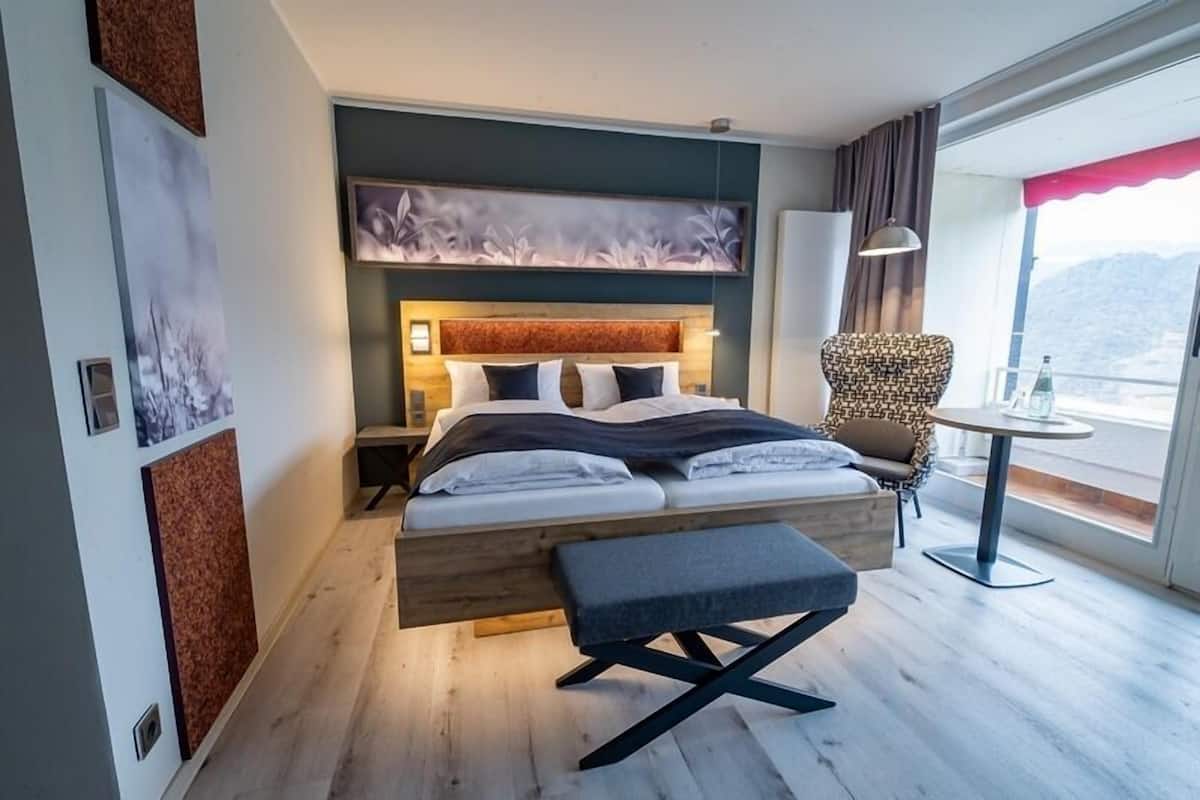 Habitación de hotel moderna con una cama grande, un banco, una silla, una mesa y vistas a las montañas.