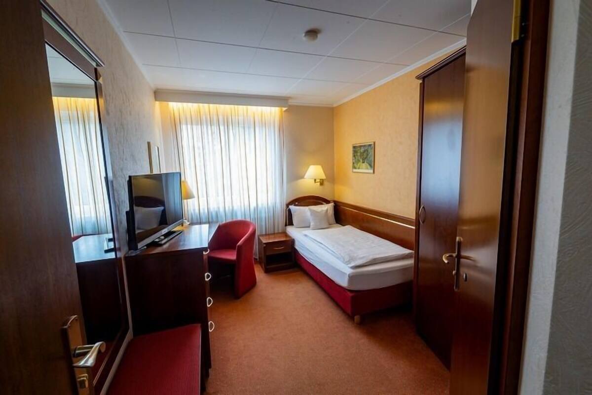Habitación de hotel con cama, escritorio con computadora, silla, mesita de noche con lámpara y una puerta.