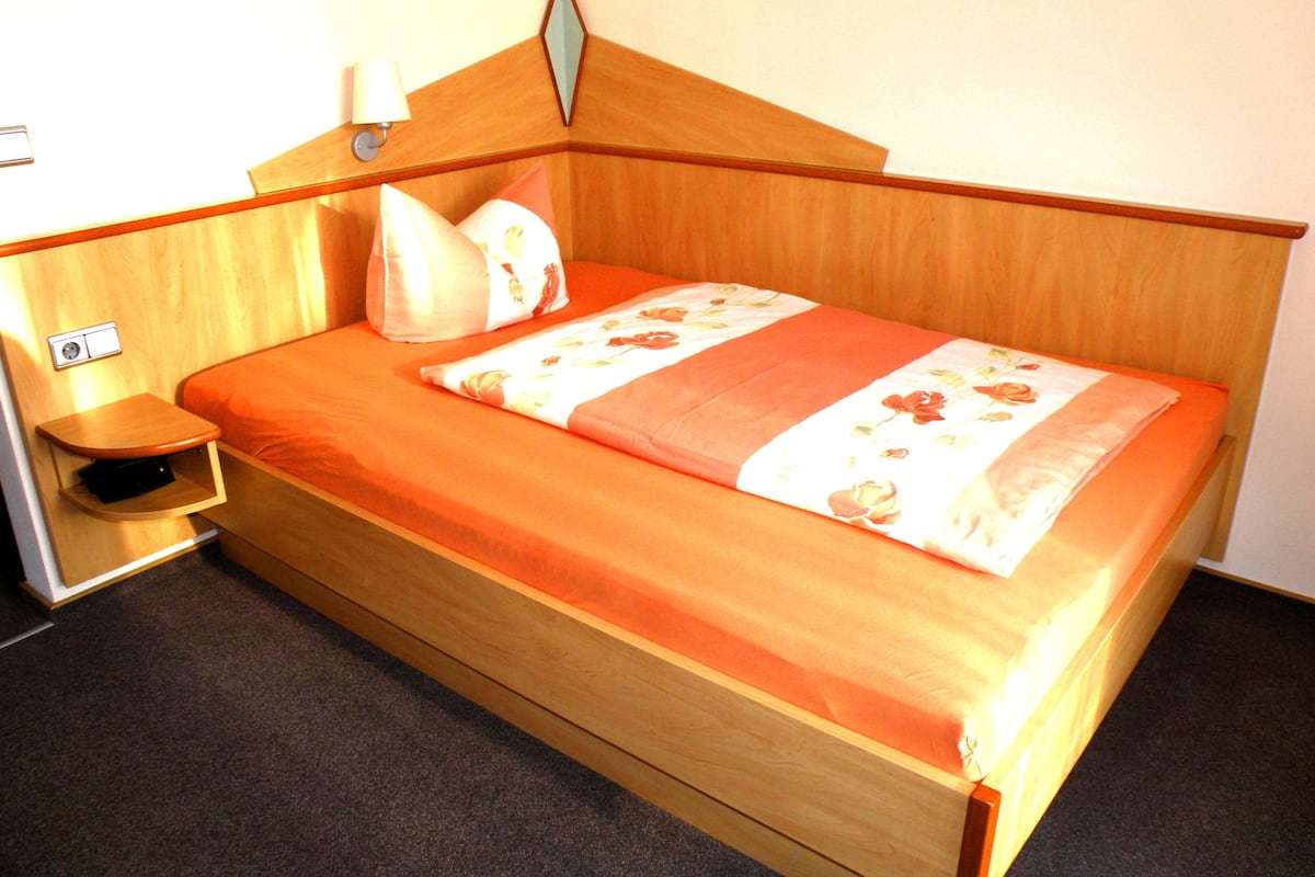A wooden bed with a floral bedspread and pillows, a bedside table, and a wall-mounted lamp.