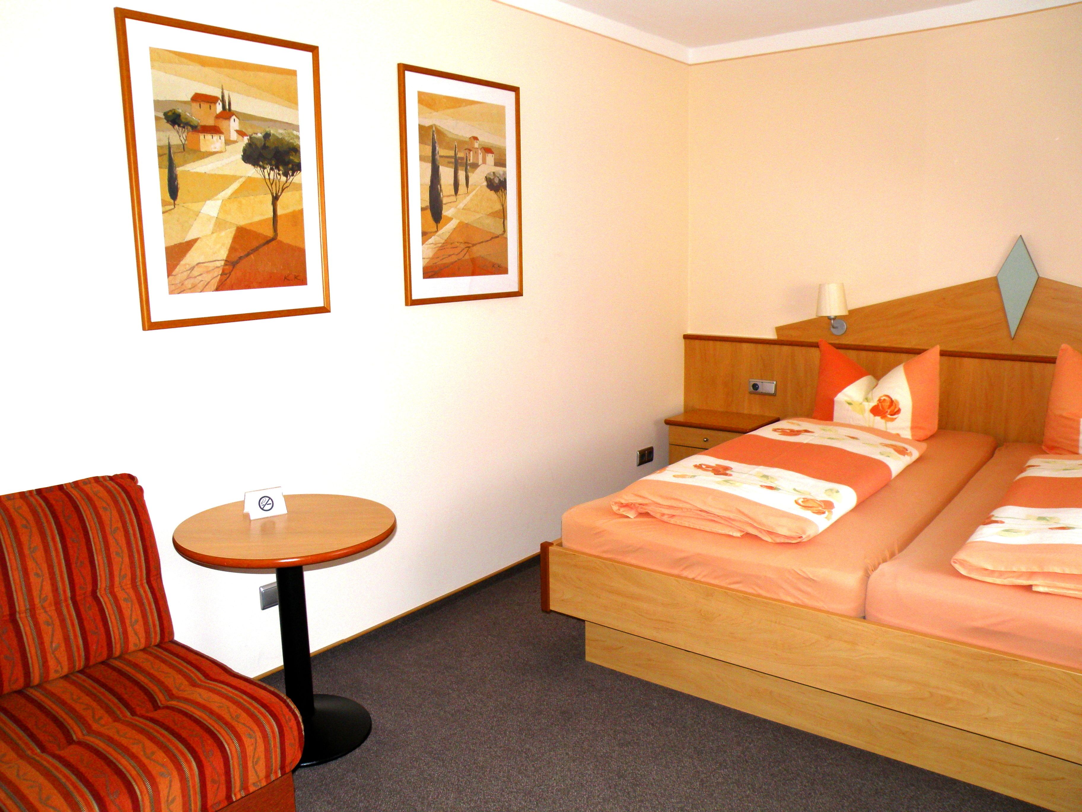 A hotel room with a bed, a chair, a small table, and two framed paintings on the wall.
