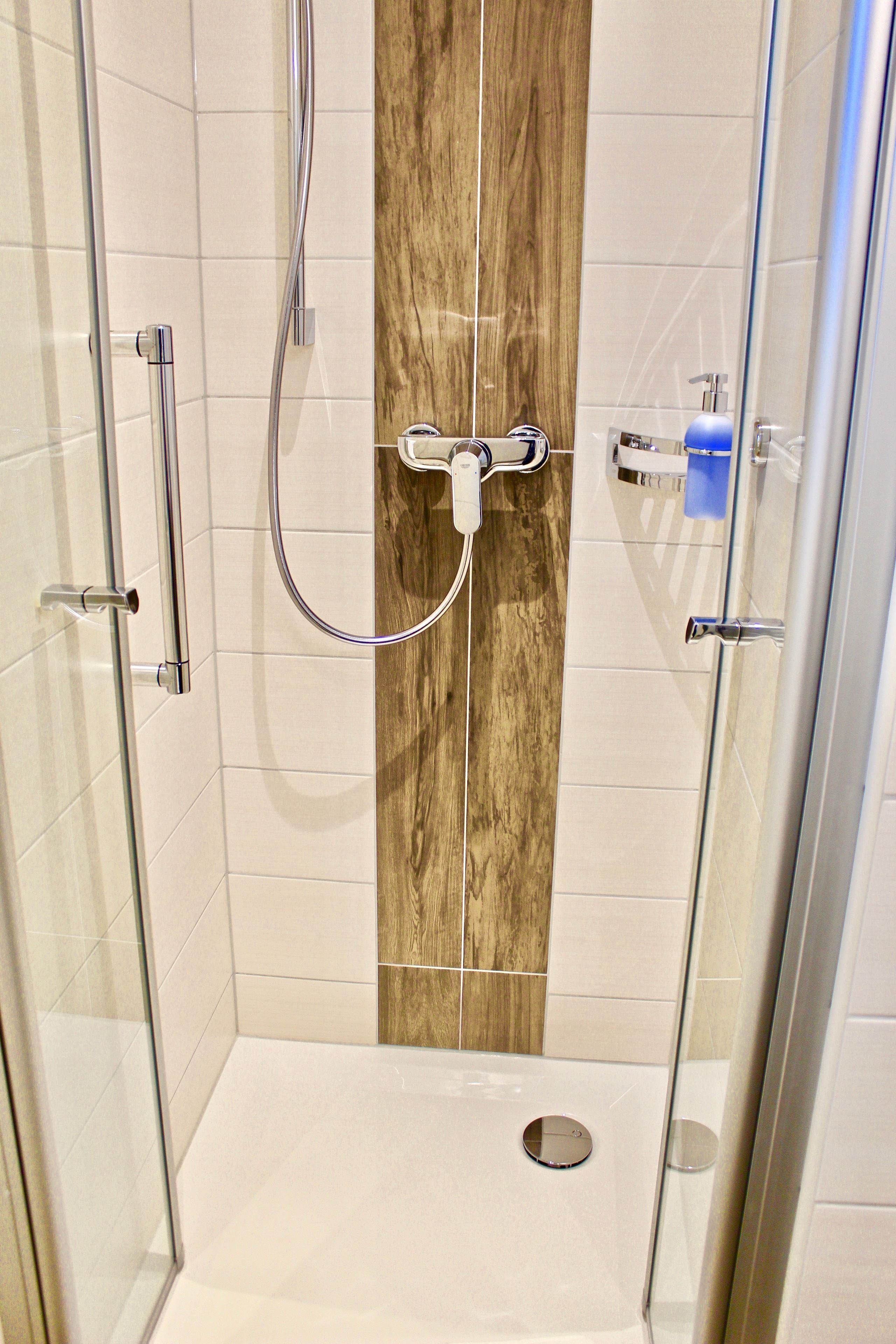 A glass-enclosed shower with a wooden accent wall, a handheld showerhead, and a control panel.