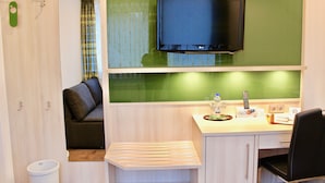 Quarto de hotel compacto com uma cama, uma mesa com cadeira, uma televisão fixada em uma parede verde e uma pequena área de estar com um sofá.