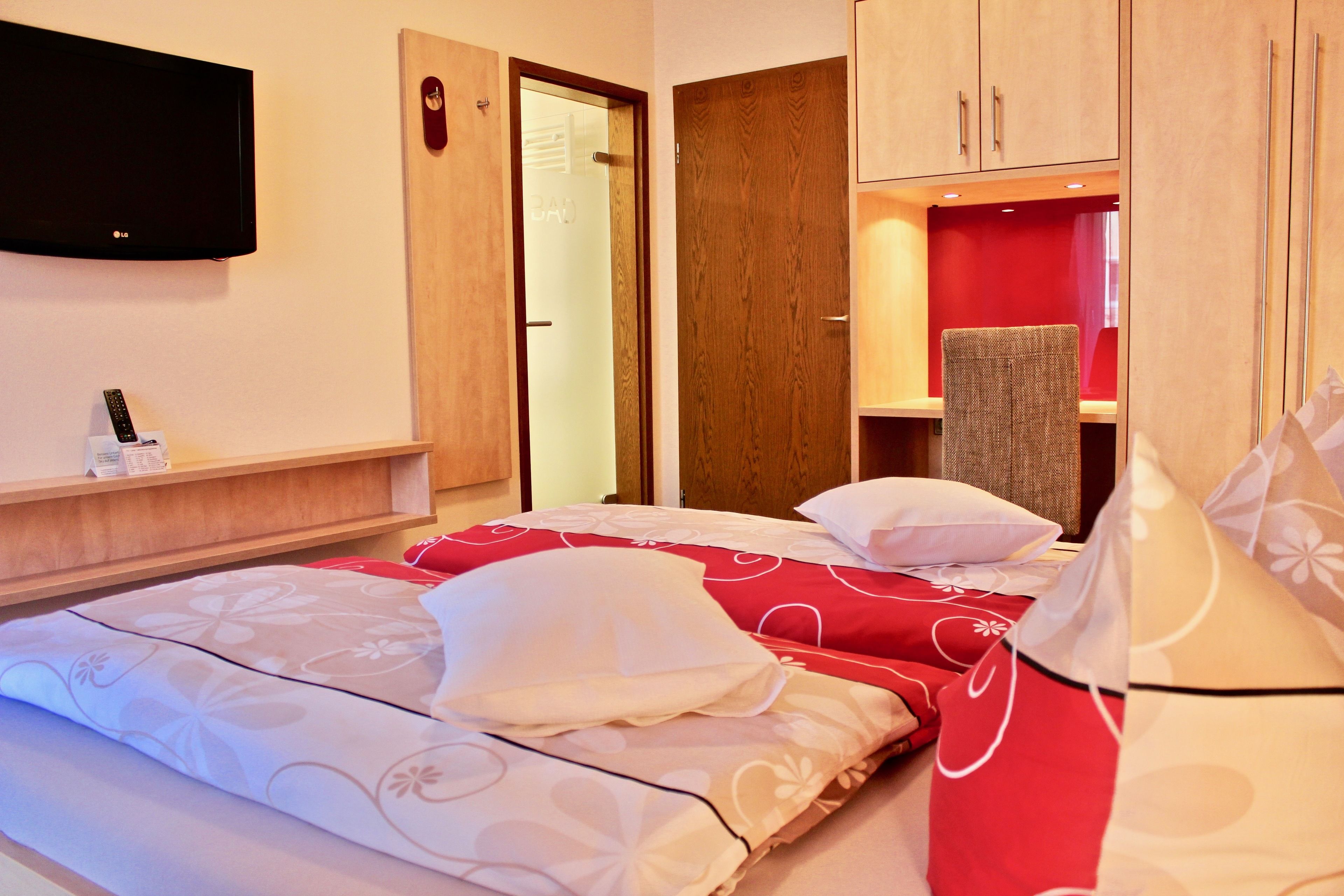 Quarto de hotel com duas camas, uma televisão, um guarda-roupa de madeira e uma parede com detalhes vermelhos.
