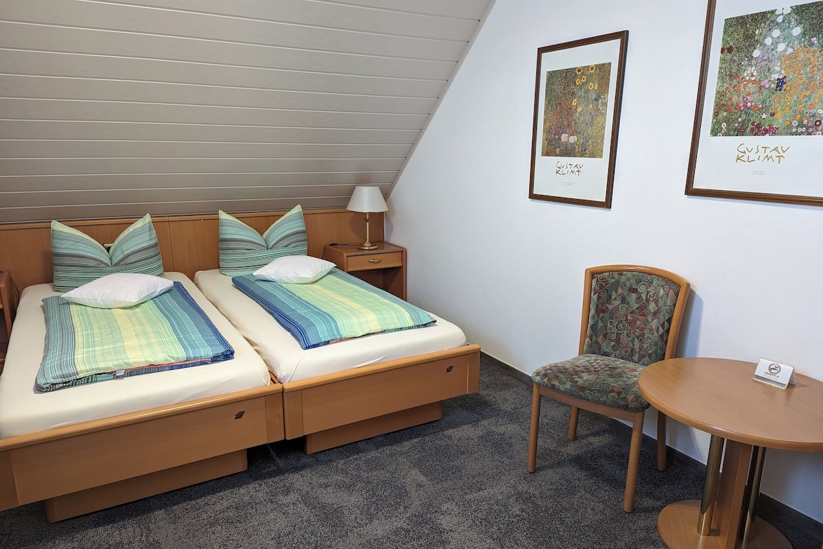 A room with two beds, a chair, a table, and two framed artworks on the wall.