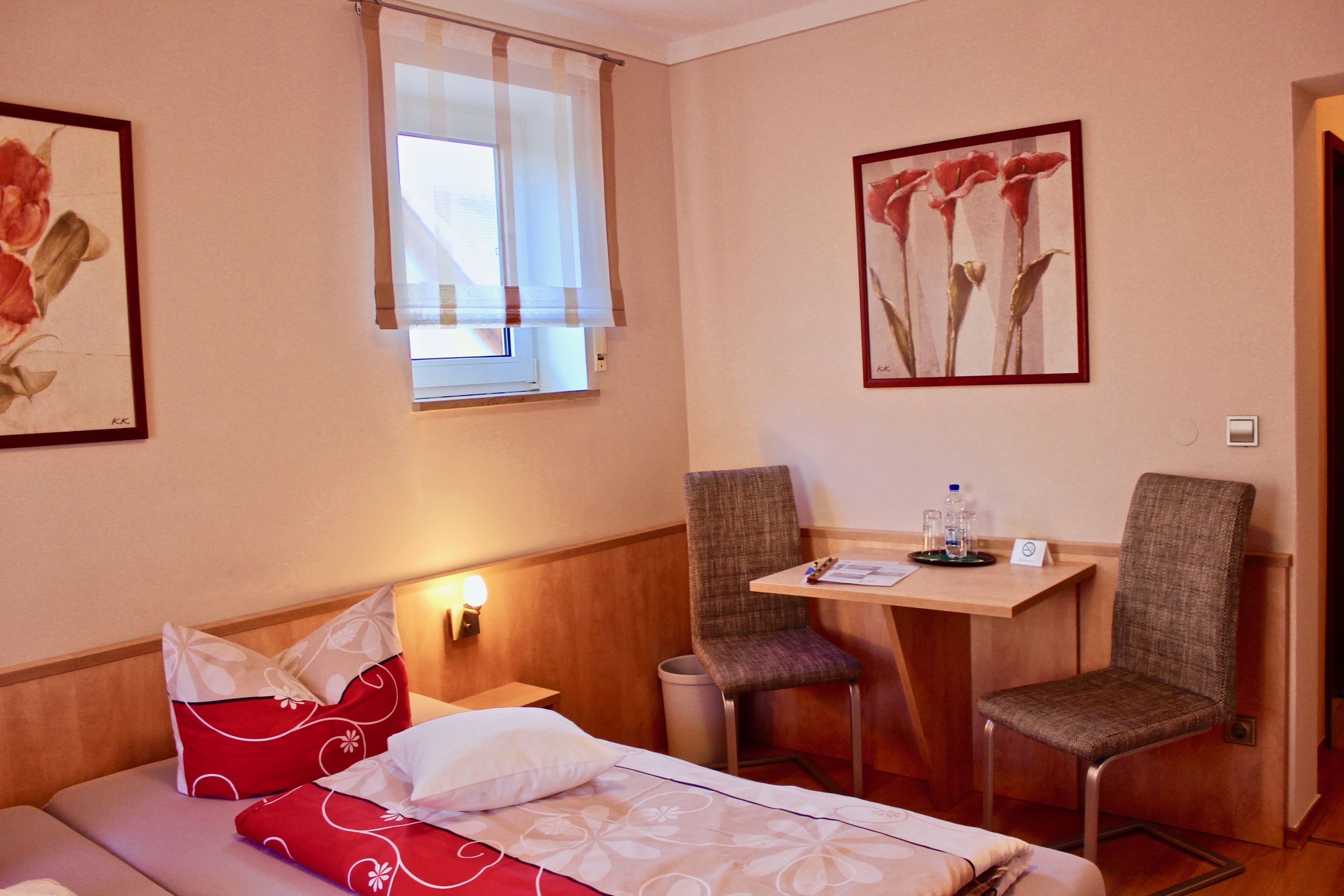Habitación de hotel con cama, dos sillas, una mesita, ventana con cortinas y un cuadro en la pared.