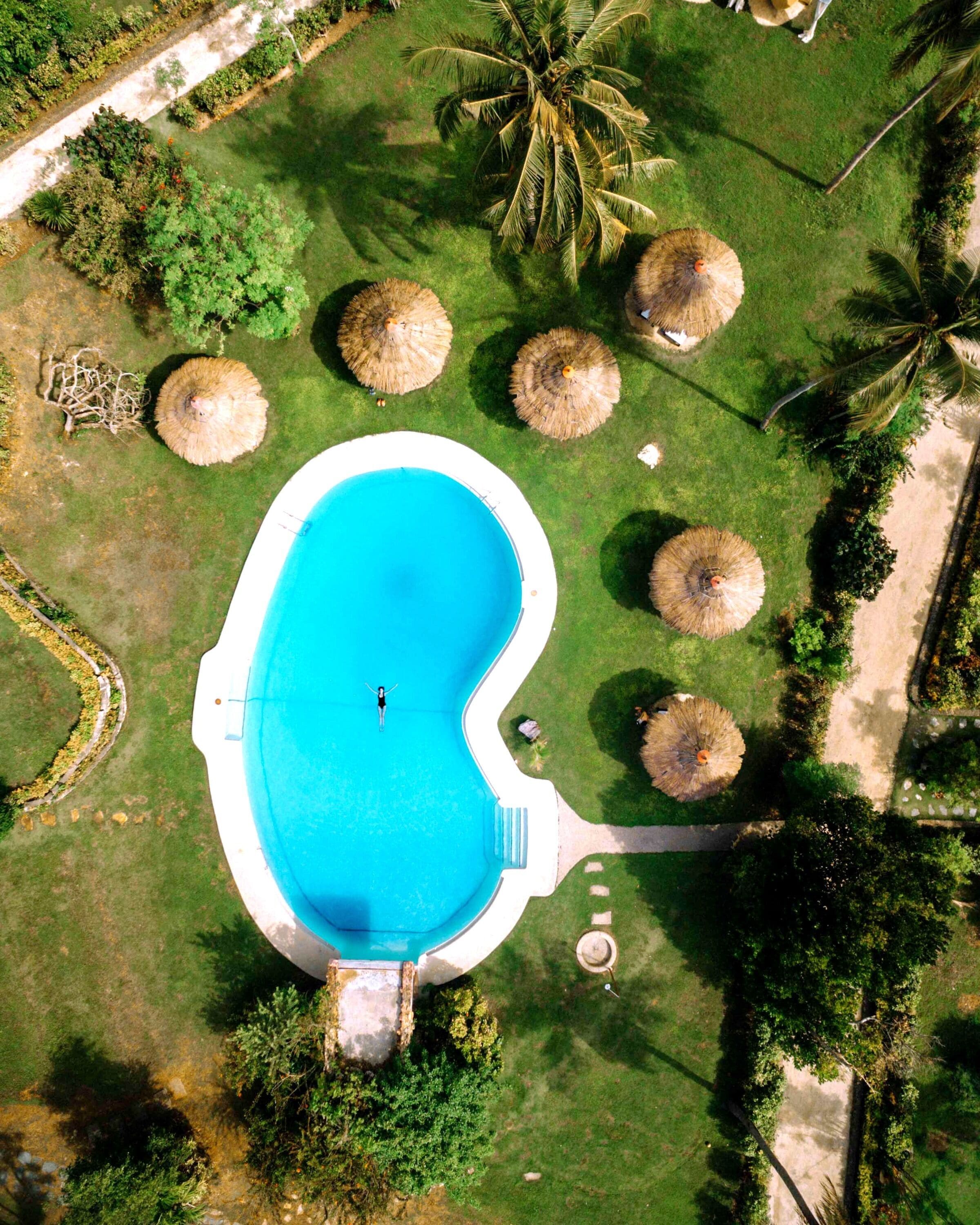 Vista aérea de una piscina rodeada de césped, palmeras y sombrillas de paja.