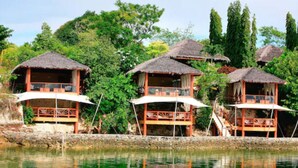 Une série de bungalows aux toits de chaume, dotés de balcons, situés au bord d’un plan d’eau et entourés d’une végétation luxuriante.