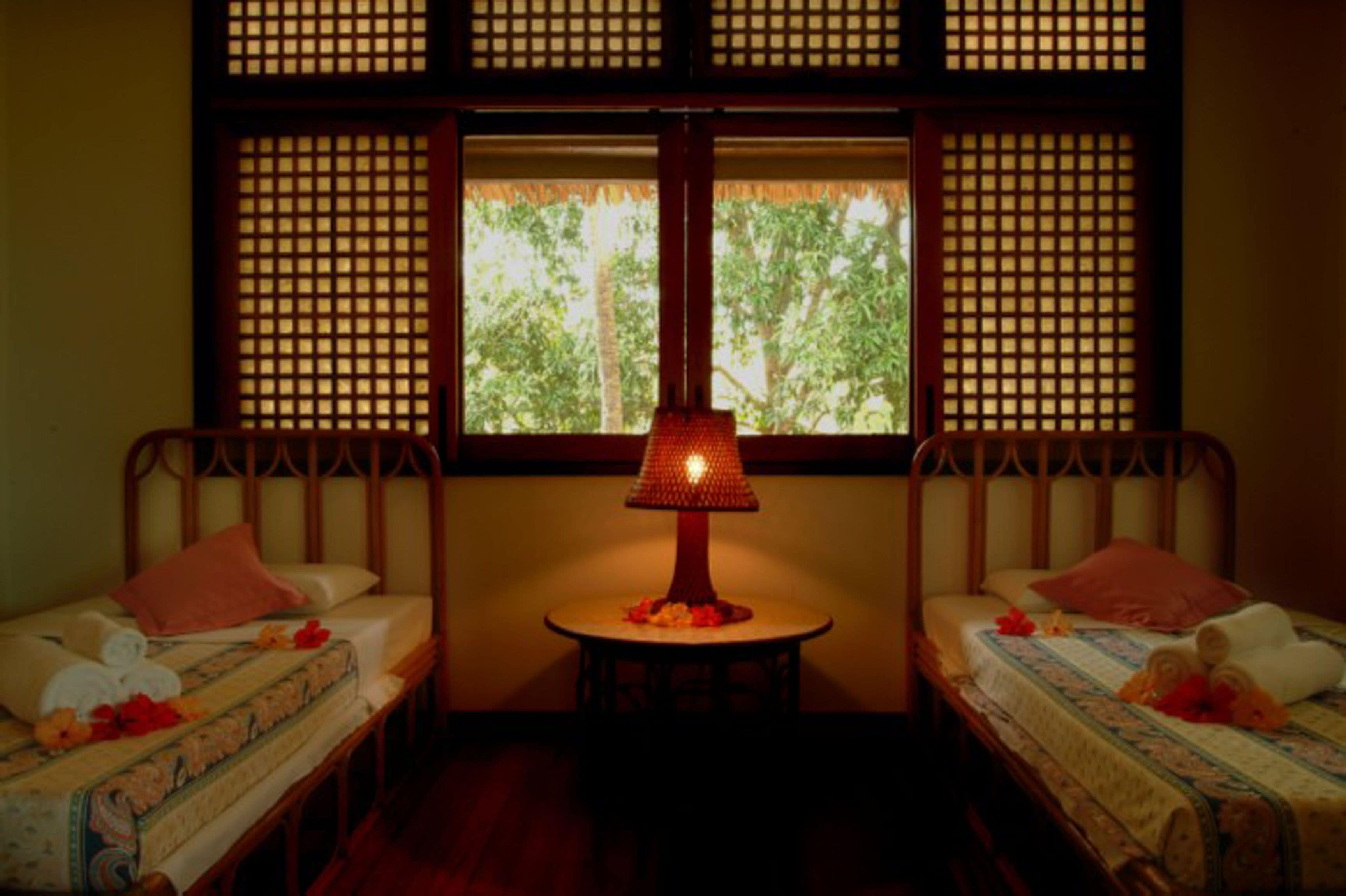 Una habitación con dos camas, una mesita con una lámpara y una ventana con vistas a árboles.