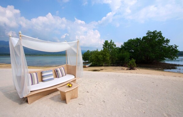 A beach setting with a wicker sofa under a canopy, a small table with a flower, and a view of the sea and trees. - Badian Island Wellness Resort (Badian)