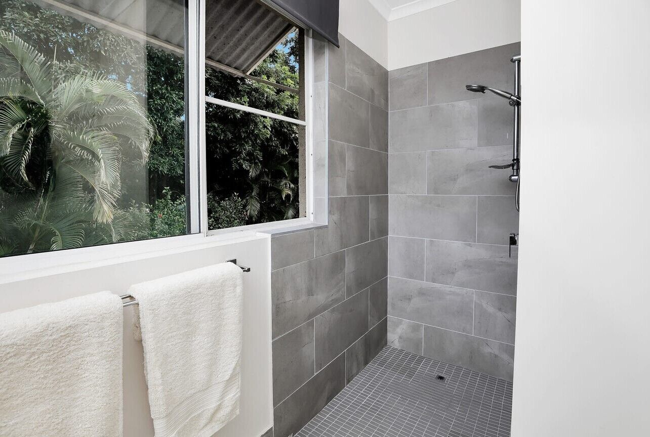 Uma casa de banho moderna com azulejos cinzentos, cabine de duche em vidro e janela com vista para vegetação.