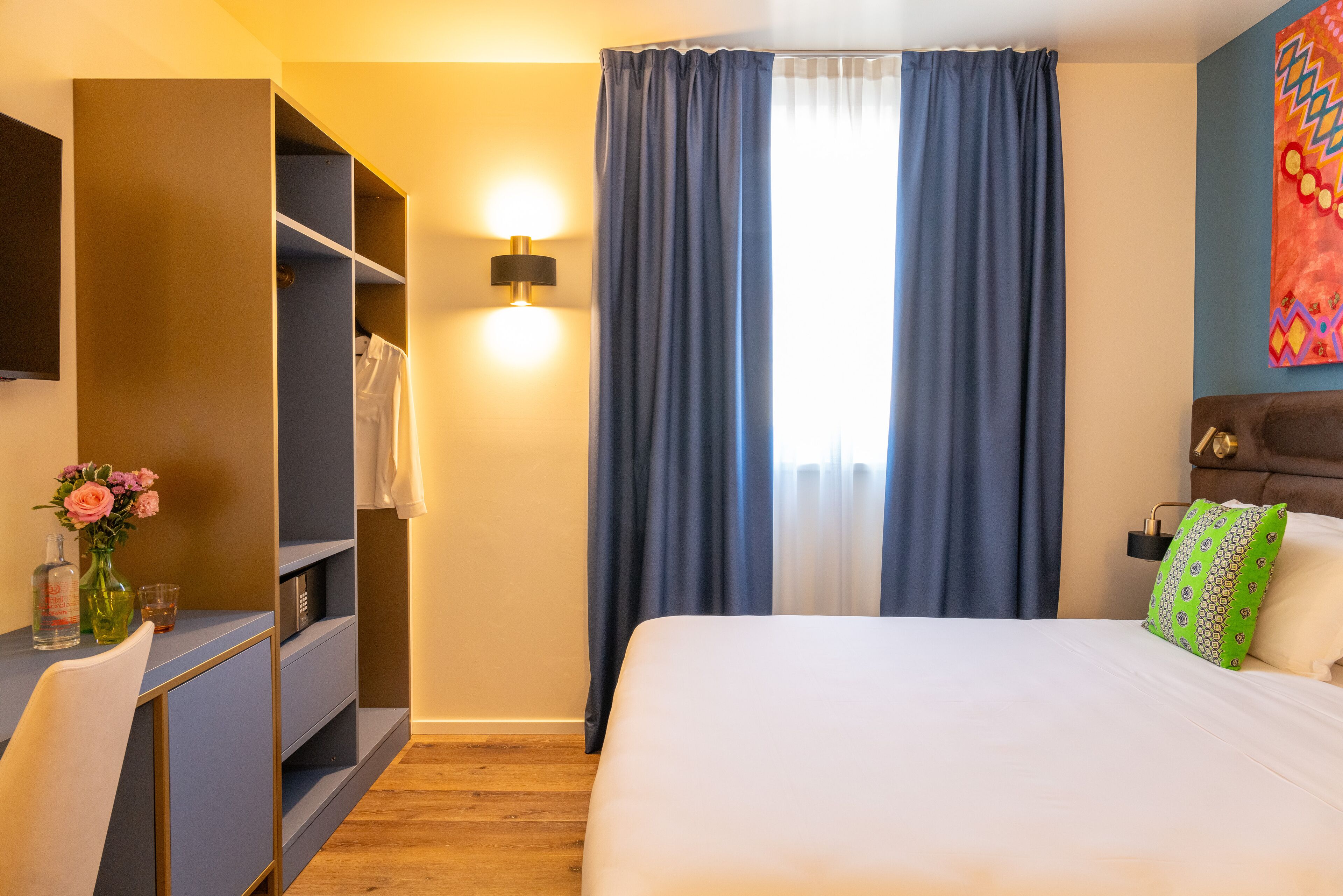 A hotel room with a bed, a desk with a chair, a wardrobe, and a wall-mounted lamp.