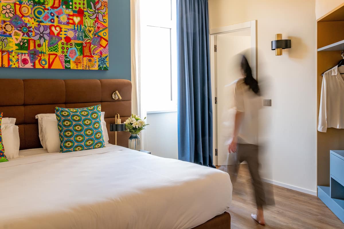 A hotel room with a large bed, a colorful abstract painting, a blue curtain, and a wardrobe.