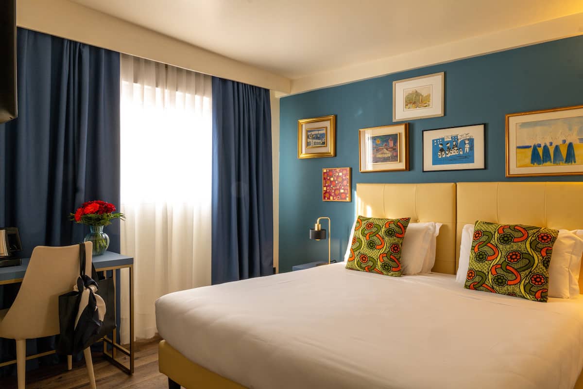 A hotel room with a bed, a desk, and a chair. There are curtains, a vase with flowers, and framed pictures on the wall.
