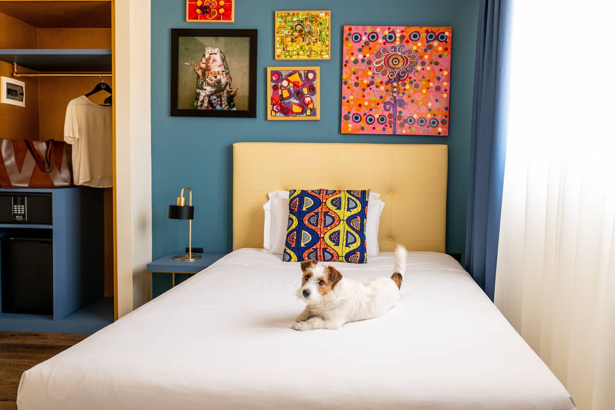 A hotel room with a bed, a small table with a lamp, a wardrobe, and artwork on the wall.