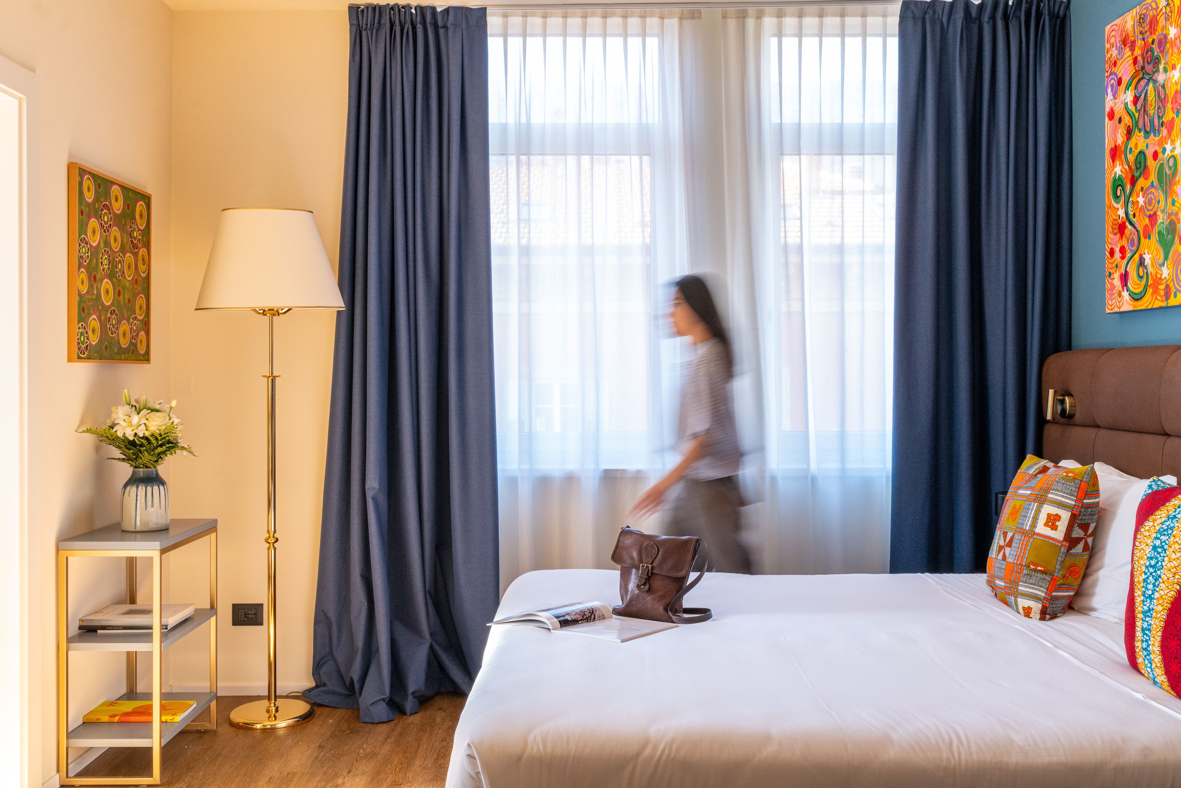 Habitación de hotel con cama, mesita de noche con lámpara, ventana con cortinas y un cuadro en la pared.