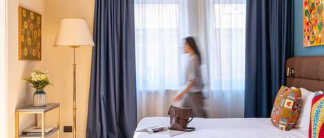 Habitación de hotel con cama, mesita de noche con lámpara, ventana con cortinas y un cuadro en la pared.