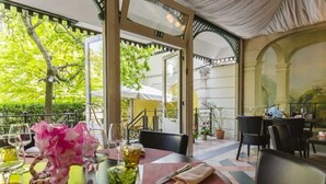 Un área de comedor al aire libre con mesas dispuestas para dos personas, con vistas a zonas verdes y un edificio al fondo.