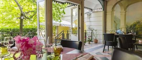 An outdoor dining area with tables set for two, featuring a view of greenery and a building in the background.