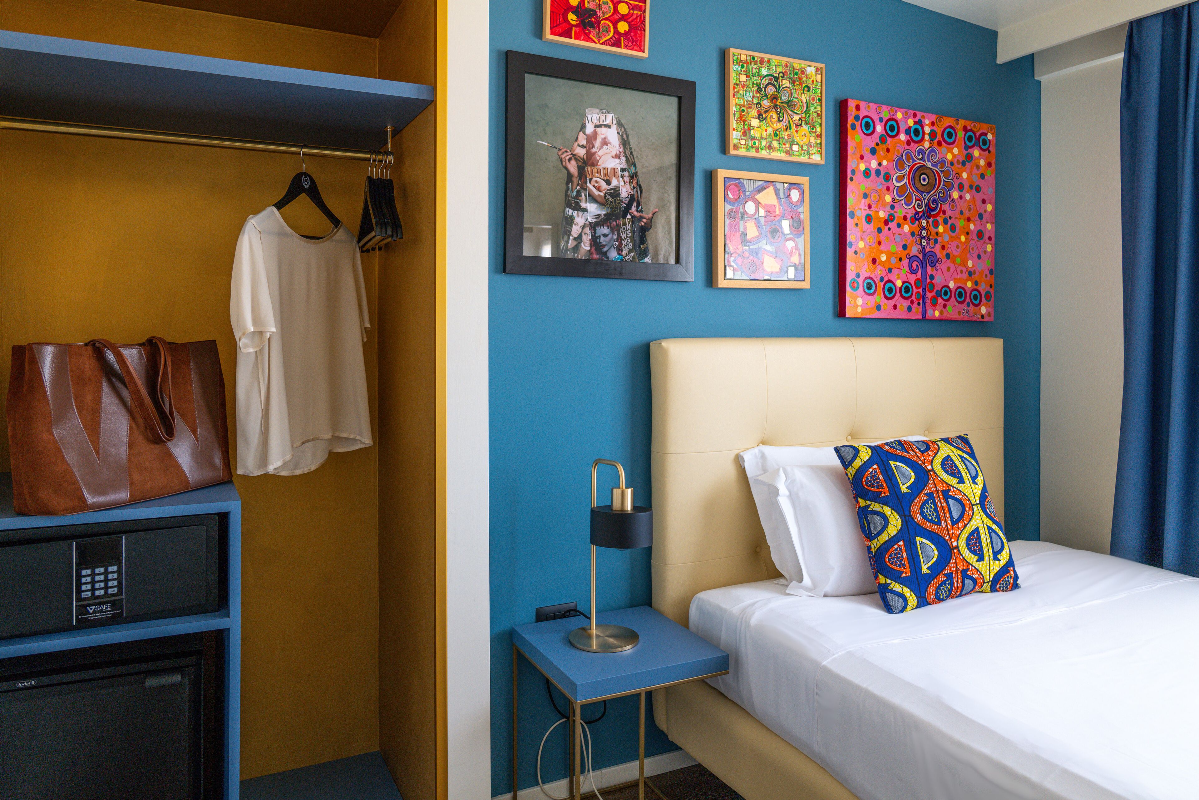 Quarto de hotel com uma cama, mesa de cabeceira, escrivaninha com computador, guarda-roupa e obras de arte nas paredes.