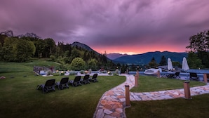 A stone pathway leading to a seating area with chairs and umbrellas, surrounded by lush greenery and mountains in the background. - Alm- & Wellnesshotel Alpenhof (Schoenau am Koenigssee)