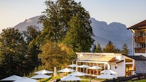 A modern building with a glass facade, outdoor seating area with umbrellas, and a view of mountains in the background. - Alm- & Wellnesshotel Alpenhof (Schoenau am Koenigssee)