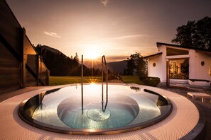 Ein Whirlpool mit kreisförmigem Design, umgeben von einem strukturierten Deck, vor einer Kulisse aus Sonnenuntergang und Berglandschaft.