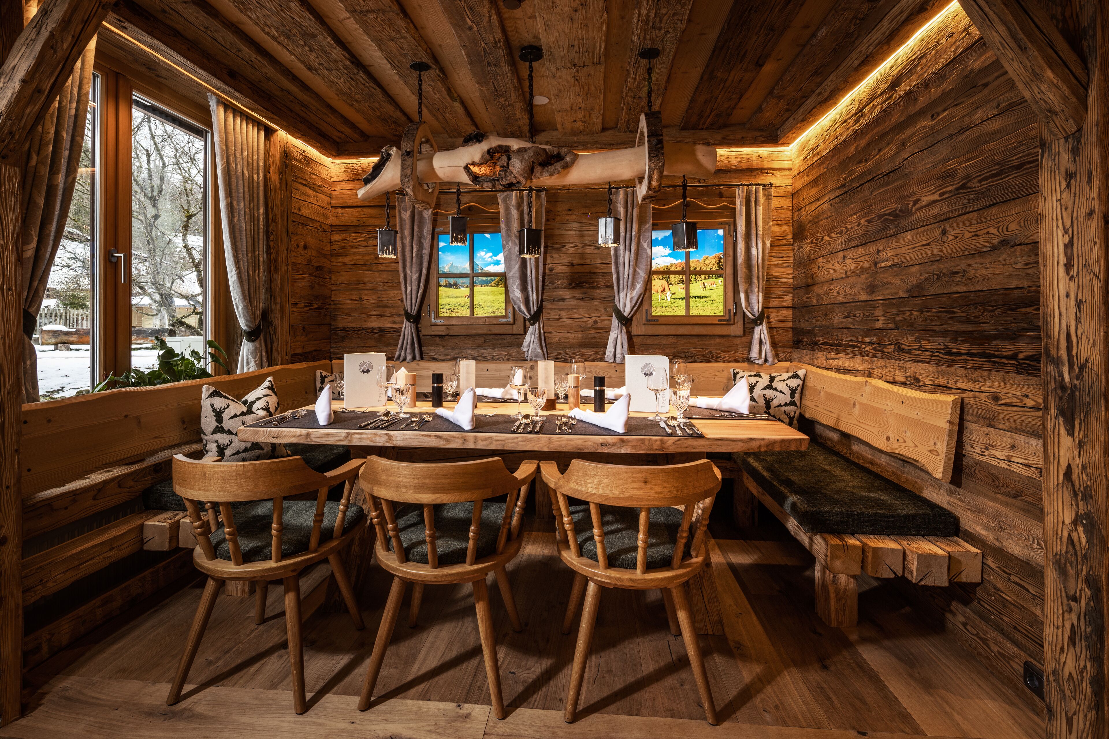 A wooden cabin interior with a dining table set for four, wooden benches, and a large window with curtains.