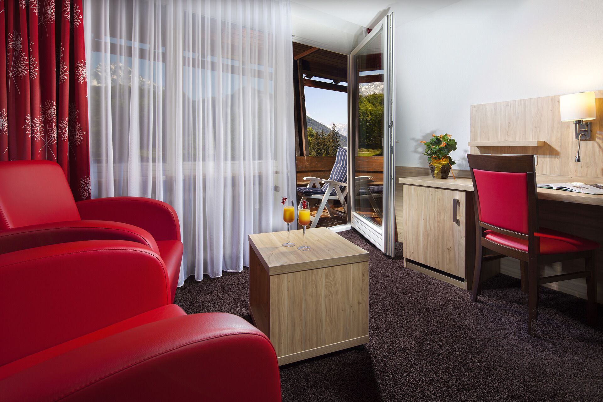 Ein Hotelzimmer mit einem roten Sofa, einer Holzkabine, einem Esstisch mit einem Stuhl und Blick auf Berge durch das Fenster.