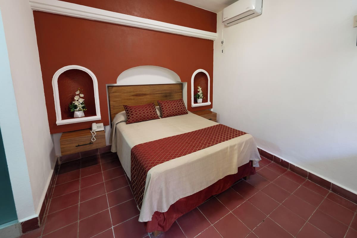 Un dormitorio con una cama, dos mesitas de noche, aire acondicionado montado en la pared y suelo de baldosas rojas.
