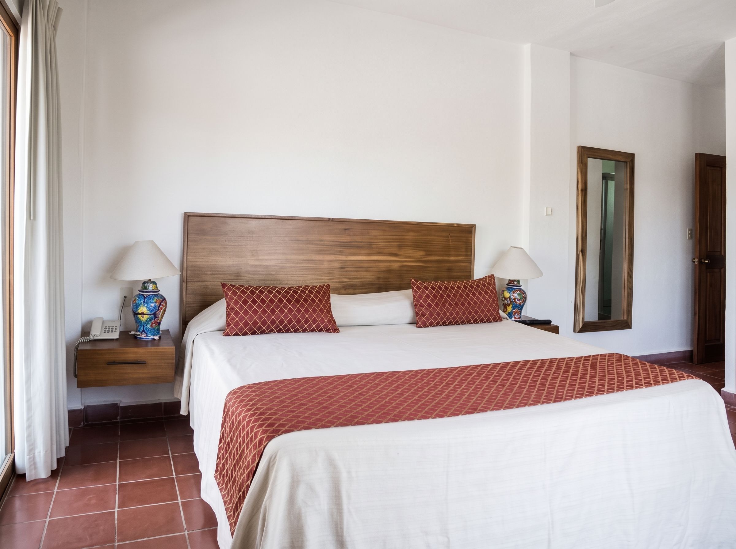 Quarto de hotel com cama grande, duas mesas de cabeceira, um criado-mudo, um espelho e uma porta.