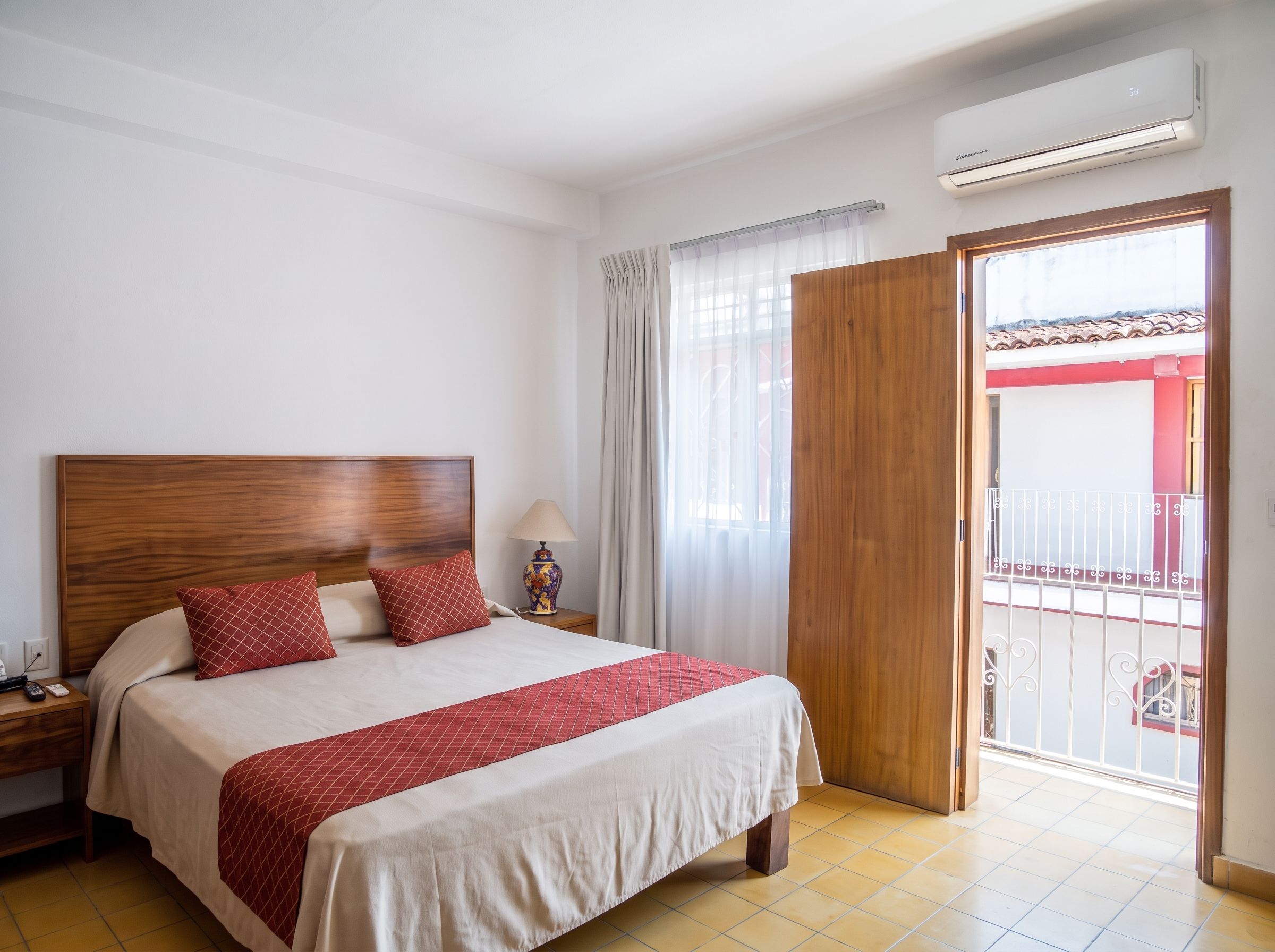 A hotel room with a wooden headboard, a bed with red and white bedding, a nightstand with a lamp, and a balcony with a red railing.