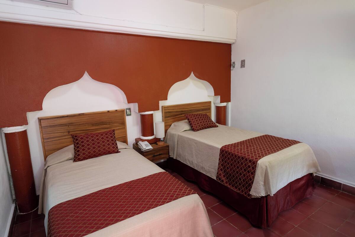 Habitación de hotel con dos camas, cada una con cabecero de madera y edredón rojo y blanco. Entre las camas, una mesita de noche con lámpara y teléfono.