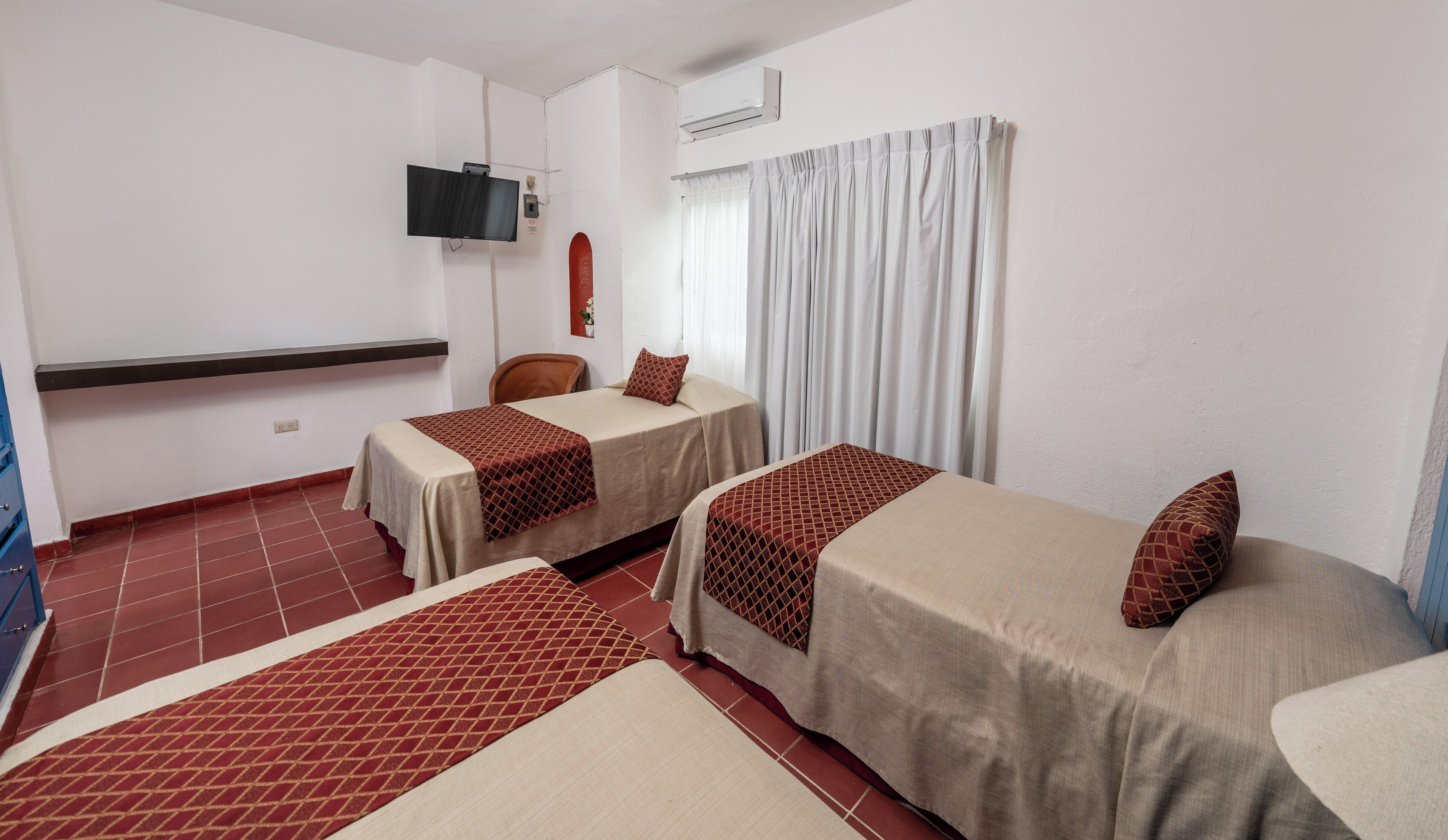 A hotel room with two single beds, a TV mounted on the wall, an air conditioning unit, and a window with curtains.