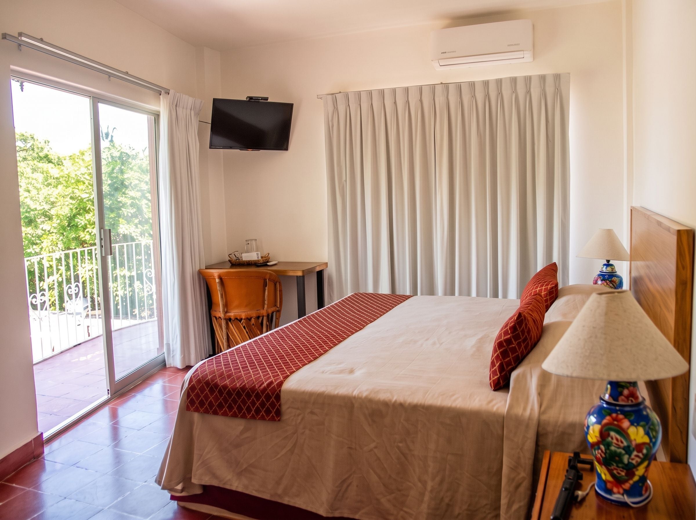 A hotel room with a bed, a table, a chair, a TV mounted on the wall, and a balcony with a view of greenery.