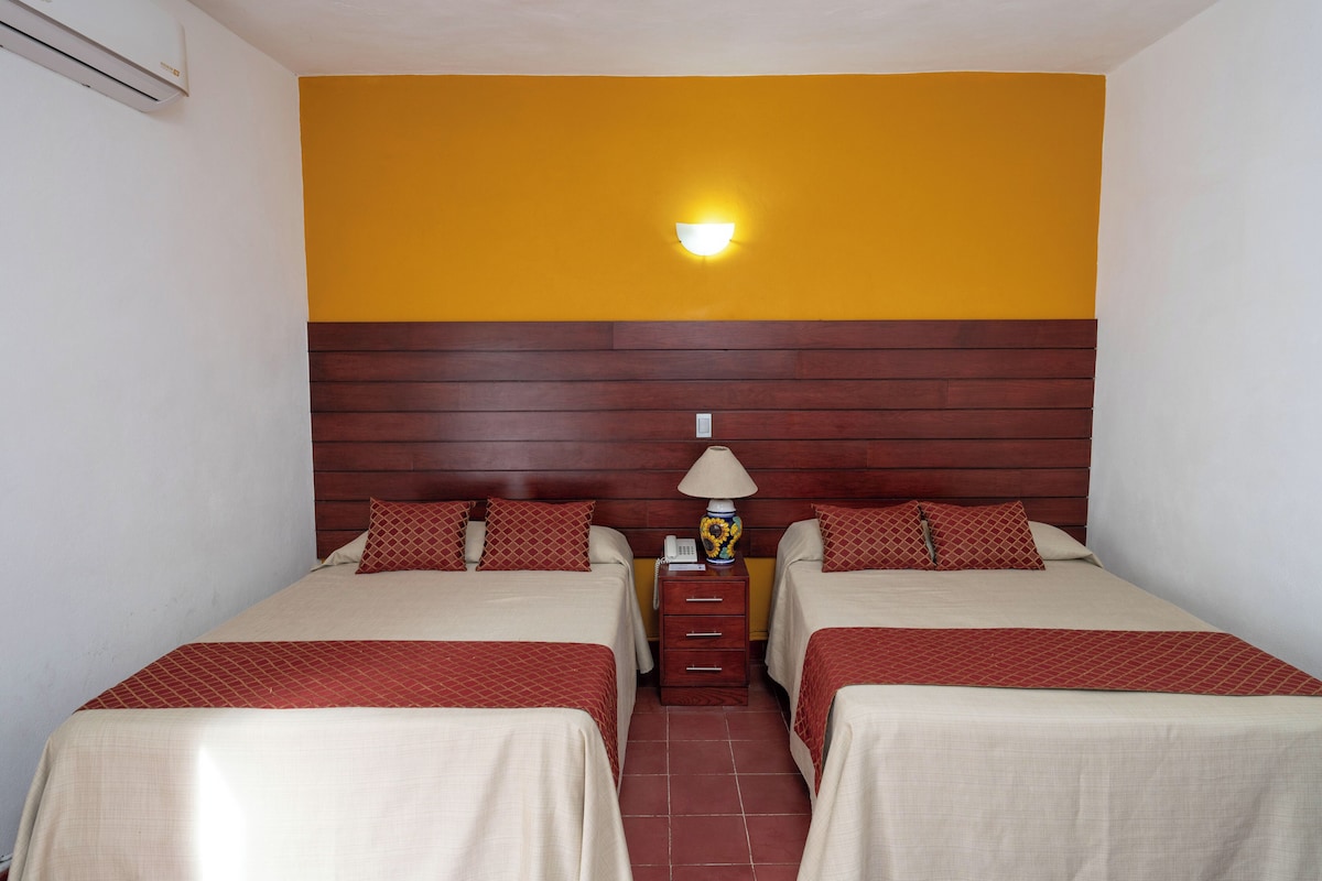 A hotel room with two beds, a wooden headboard, a nightstand with a lamp, and a wall-mounted light fixture.