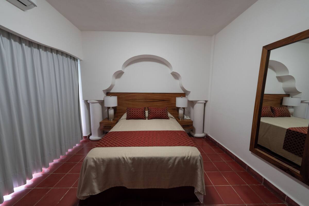 Habitación de hotel con cama, mesitas de noche, lámparas, un espejo y una ventana con cortinas.