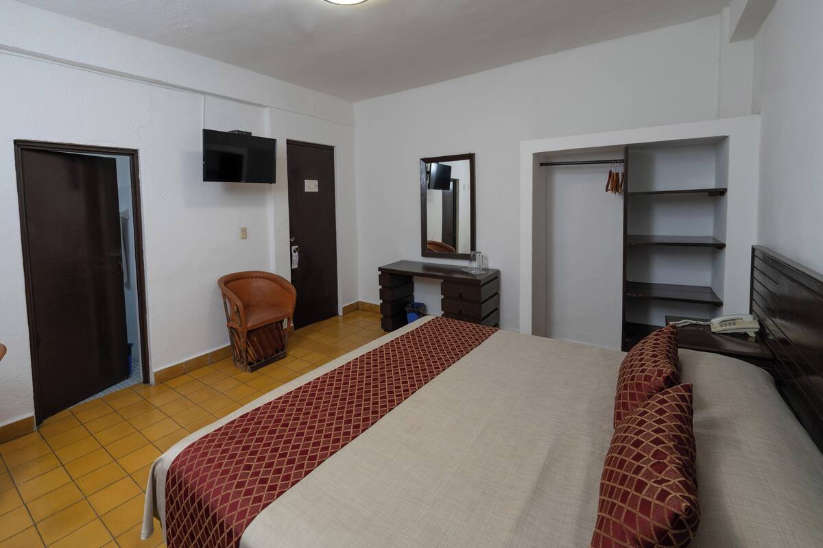 Habitación de hotel con cama, una silla, escritorio, espejo, televisor y armario.