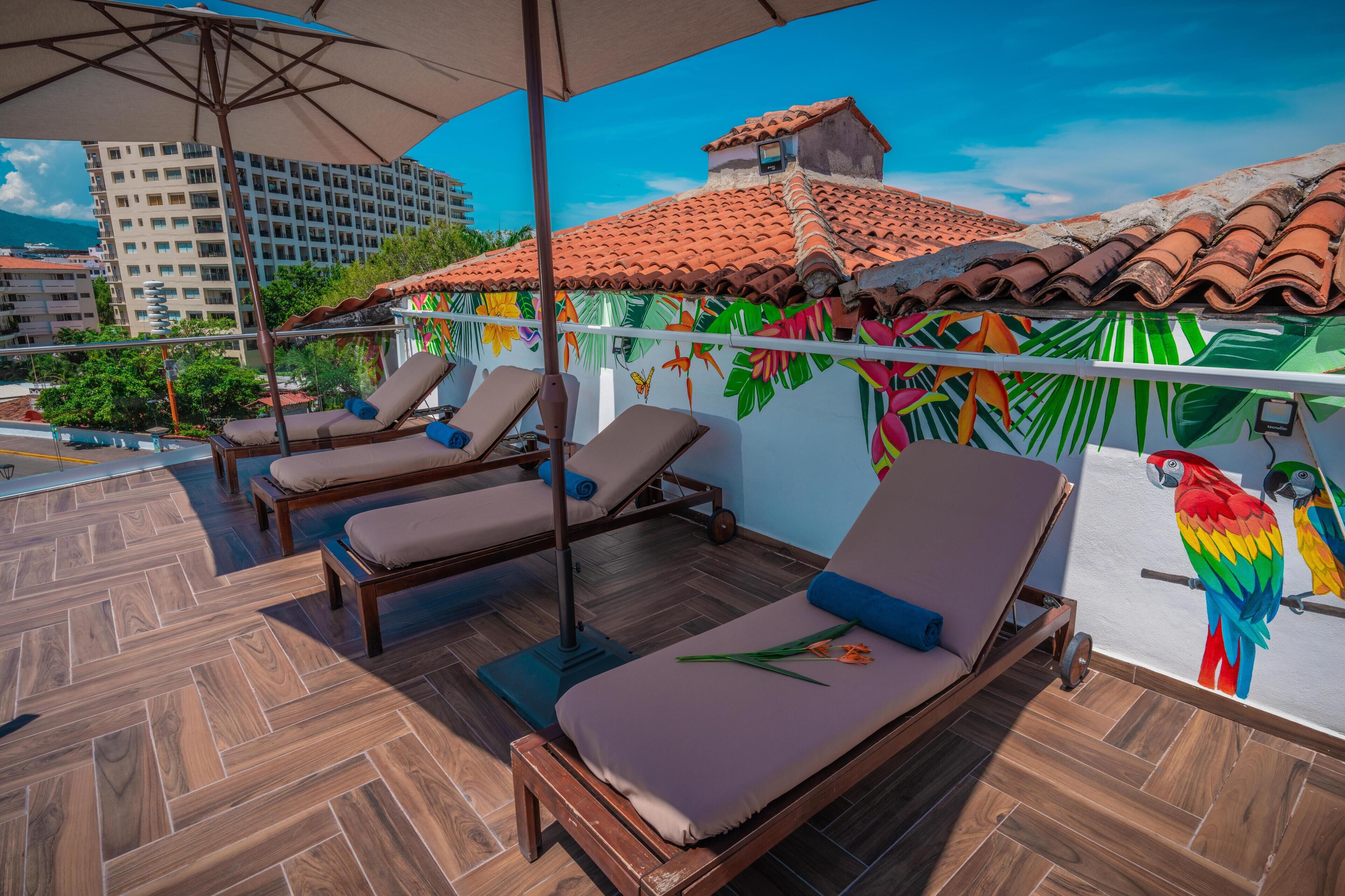 A rooftop lounge area with wooden flooring, lounge chairs, and a mural of a building and parrots.
