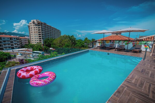 A swimming pool with two pink inflatable rings, surrounded by lounge chairs and umbrellas, with buildings and greenery in the background. - Hotel Encino Malecón Centro Puerto Vallarta (Puerto Vallarta)