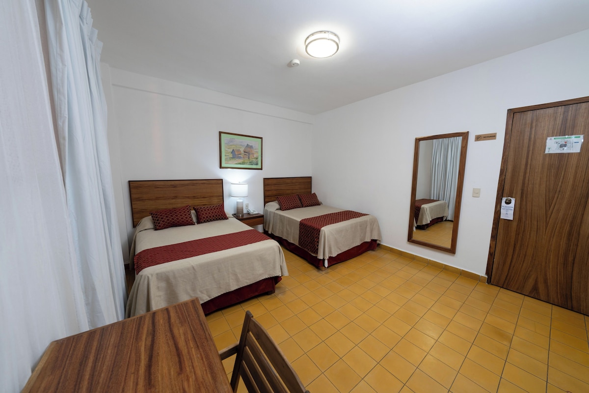A hotel room with two beds, a wooden table, a chair, a mirror, and a ceiling light.