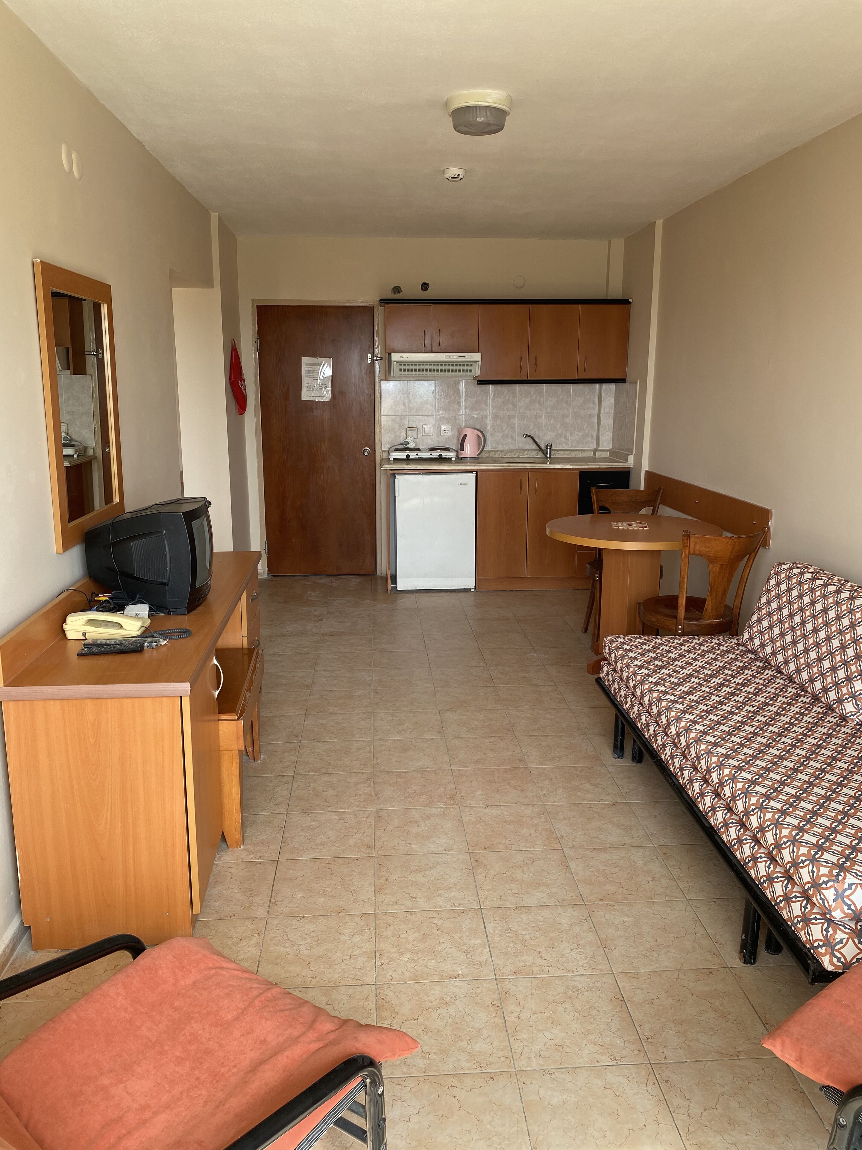 A compact room with a kitchenette, a small dining table, a sofa, and a desk with a television and telephone.