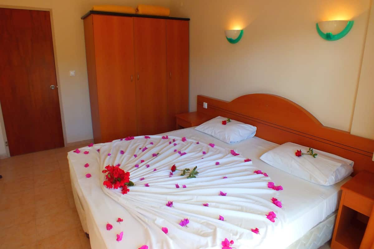 Een net opgemaakt bed met witte lakens en roze bloemblaadjes, een houten garderobe en een nachtkastje met een lamp.