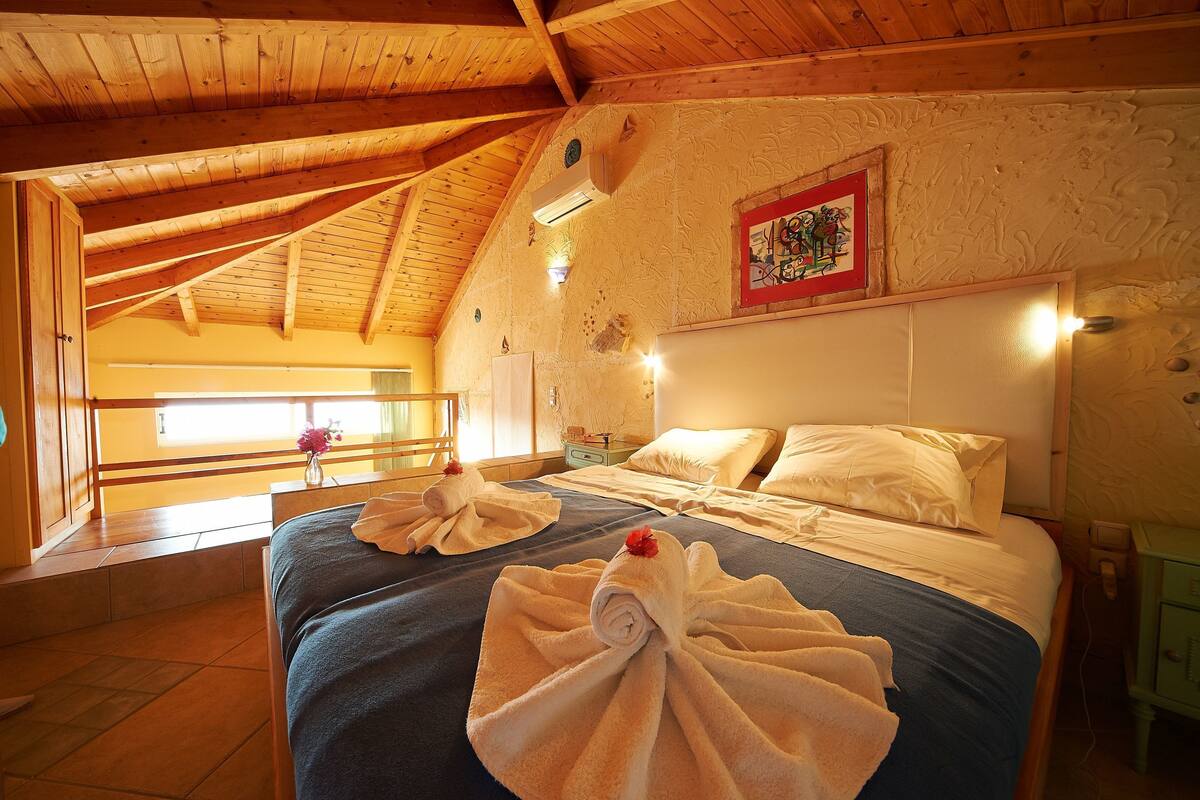 A room with a wooden ceiling, a bed with white linens, and a view through a window.