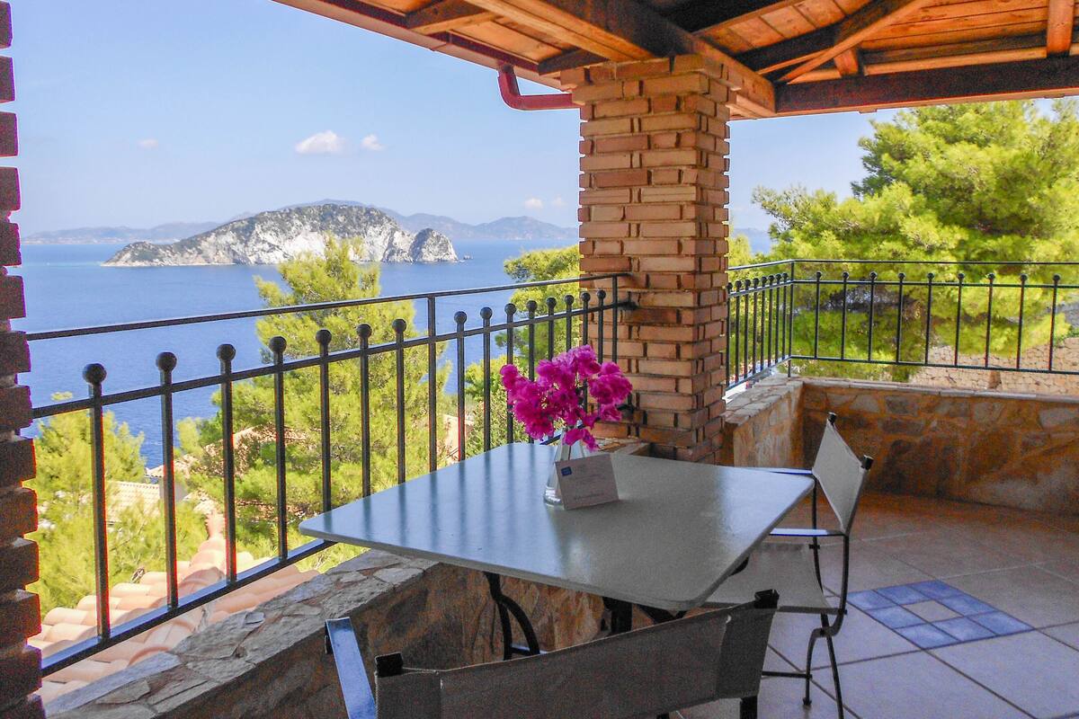 A terrace with a table and chairs, a brick pillar, and a view of the sea and a distant island.