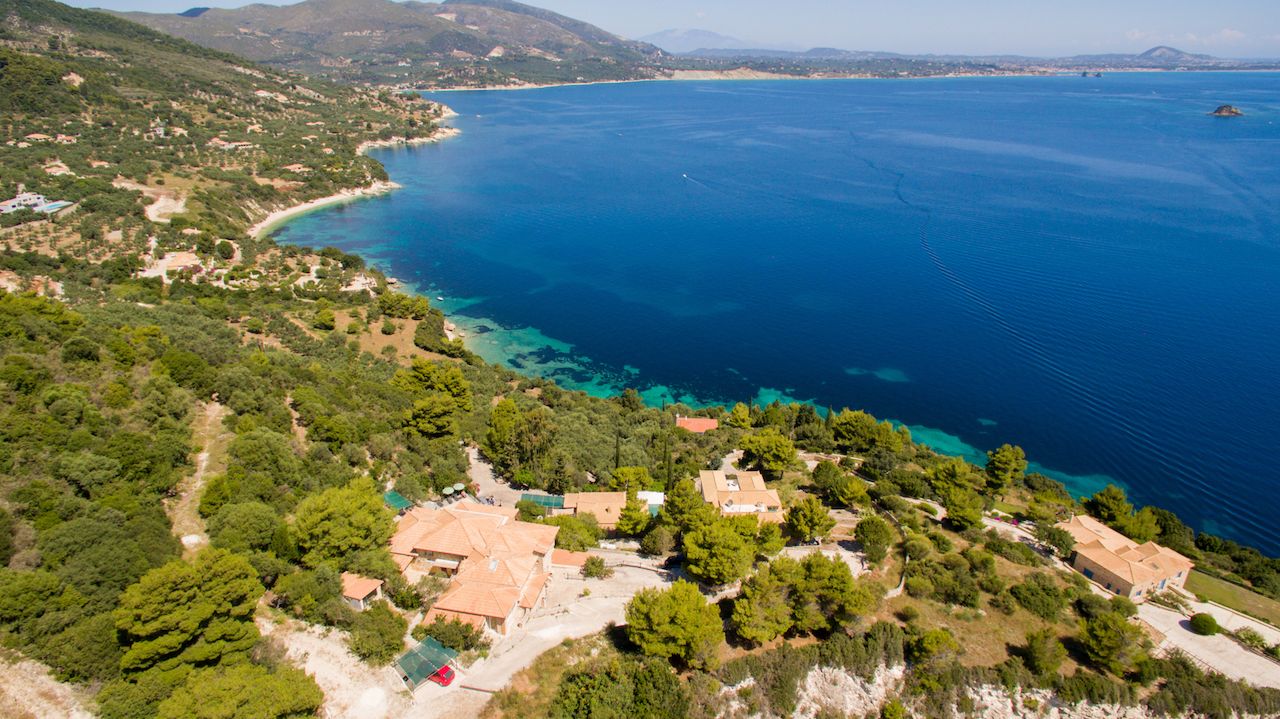 Luftaufnahme einer Küstenlandschaft mit Häusern, blauem Meer und grünen Hügeln.