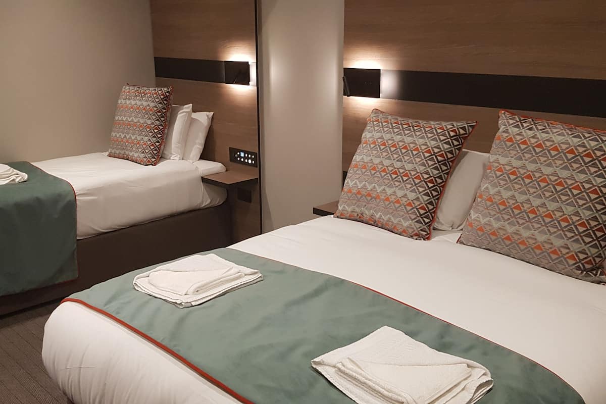 A hotel room with two beds, each with a green bedspread and white linens. There are decorative pillows on both beds, and a control panel on the wall between the beds.