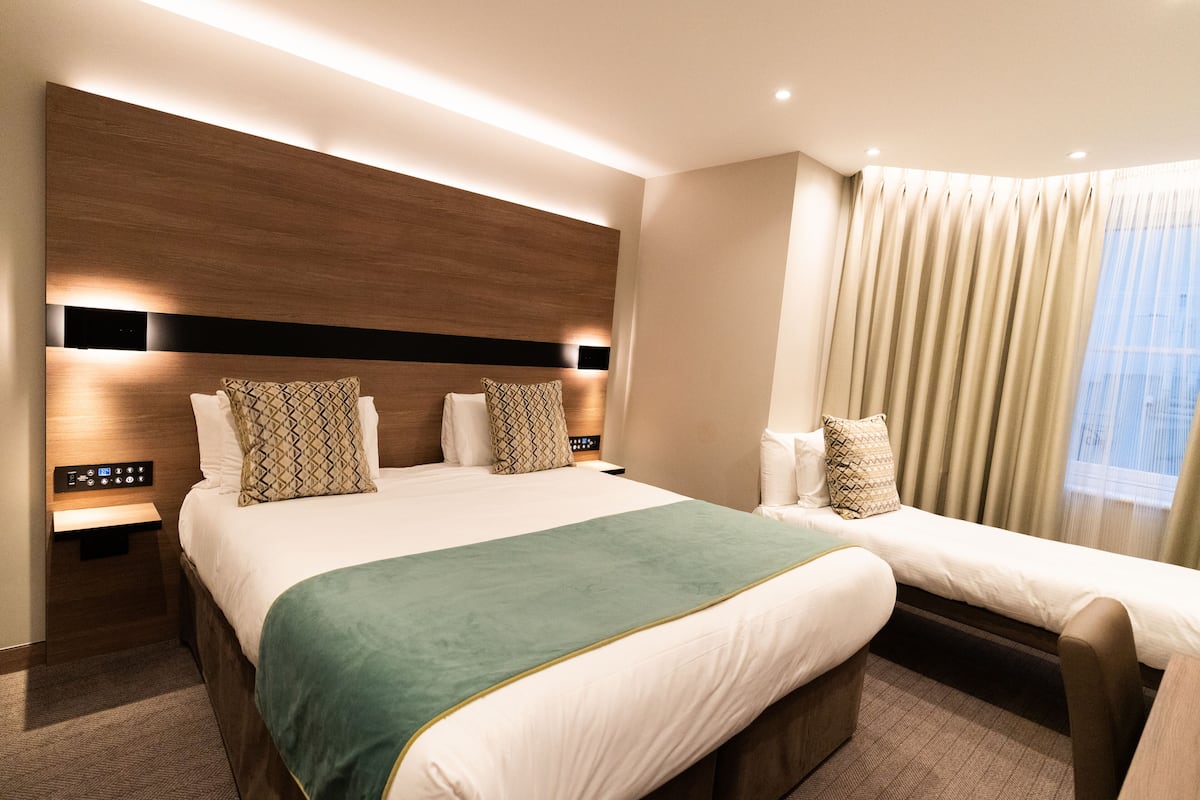 A modern hotel room with a large bed, a bedside table with a control panel, a bench with a cushion, and a large window with curtains.